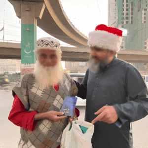Two men with white beards, one wearing a red Santa hat and the other in a patterned cap, stand under an overpass. The man in the cap holds a Gospel of John, while the other hands him a bag. Both are smiling.