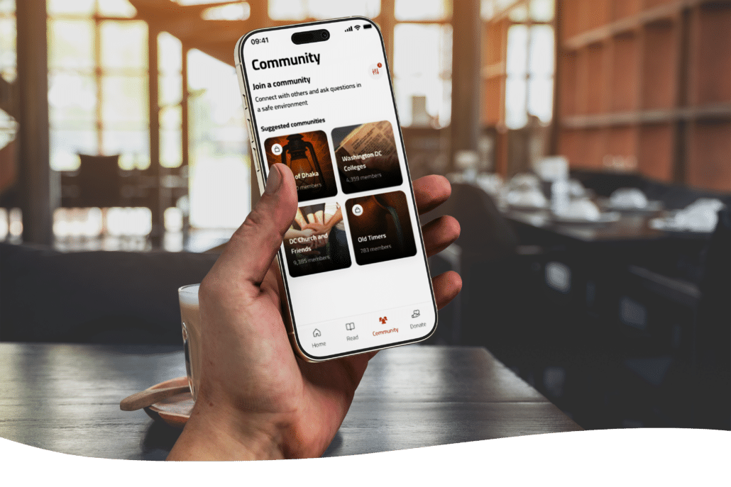 A person holds a smartphone displaying a “Community” app screen with suggested communities, sitting at a table in a warmly lit café or restaurant with blurred background.