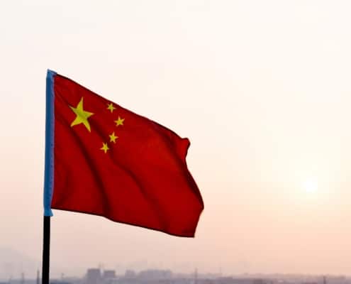 The Chinese flag waving against a hazy sky at sunset, with a cityscape visible in the background.