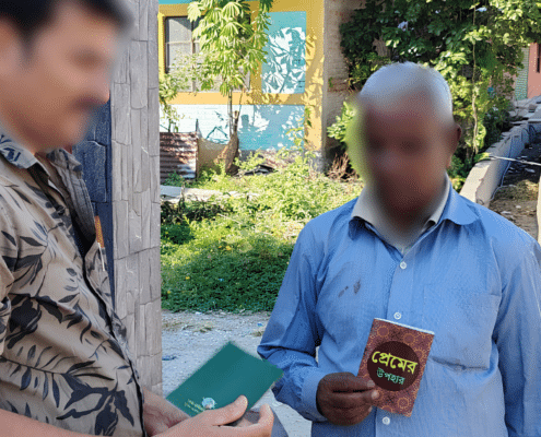 Two men with blurred faces stand outside. One holds a green book, the other holds a brown book with text in Bengali. A colorful building, greenery, and sunlight are visible in the background.
