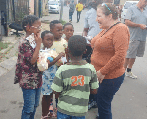 A woman in an orange top smiles while talking to a group of children and adults on a street in a residential area. The children are holding snacks, and people are gathered casually in the background.