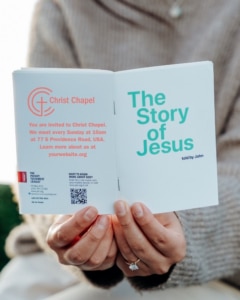 Person holding an open booklet titled "The Story of Jesus told by John" with an invitation to Christ Chapel on the left page, including meeting details and a website link.