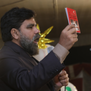 A bearded man in a suit holds up a small red book and speaks into a microphone at an indoor event with a gold star decoration in the background.