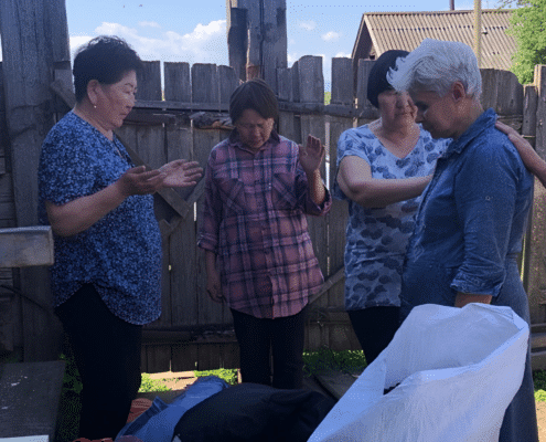 Four women stand together outdoors near a wooden fence, some with closed eyes and raised hands, appearing to pray. Bags and belongings sit in front of them. The scene is calm with sunlight and a few clouds in the sky.