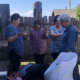 Four women stand together outdoors near a wooden fence, some with closed eyes and raised hands, appearing to pray. Bags and belongings sit in front of them. The scene is calm with sunlight and a few clouds in the sky.