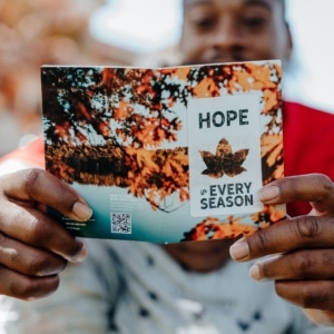 A person holds an open card with autumn leaves. The card reads “HOPE in EVERY SEASON” with a leaf graphic. The person’s face is out of focus in the background.