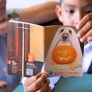 A child holds a booklet showing a dog dressed as a ghost holding a pumpkin bucket. The cover text reads, “No Tricks, Just Treats.” The booklet also displays a carved pumpkin and a QR code.