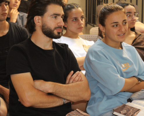 A group of people sit closely together in a room, attentively listening. Two people in front have their arms crossed and are holding brown booklets. Everyone appears focused on something out of the frame.