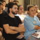 A group of people sit closely together in a room, attentively listening. Two people in front have their arms crossed and are holding brown booklets. Everyone appears focused on something out of the frame.