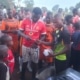 A man wearing a medal hands an item to another man, surrounded by children and adults in sports uniforms at an outdoor gathering.