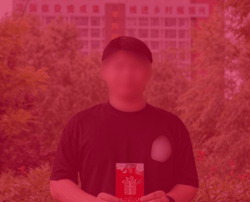A person in a black t-shirt standing in front of a building, holding a red gift envelope.
